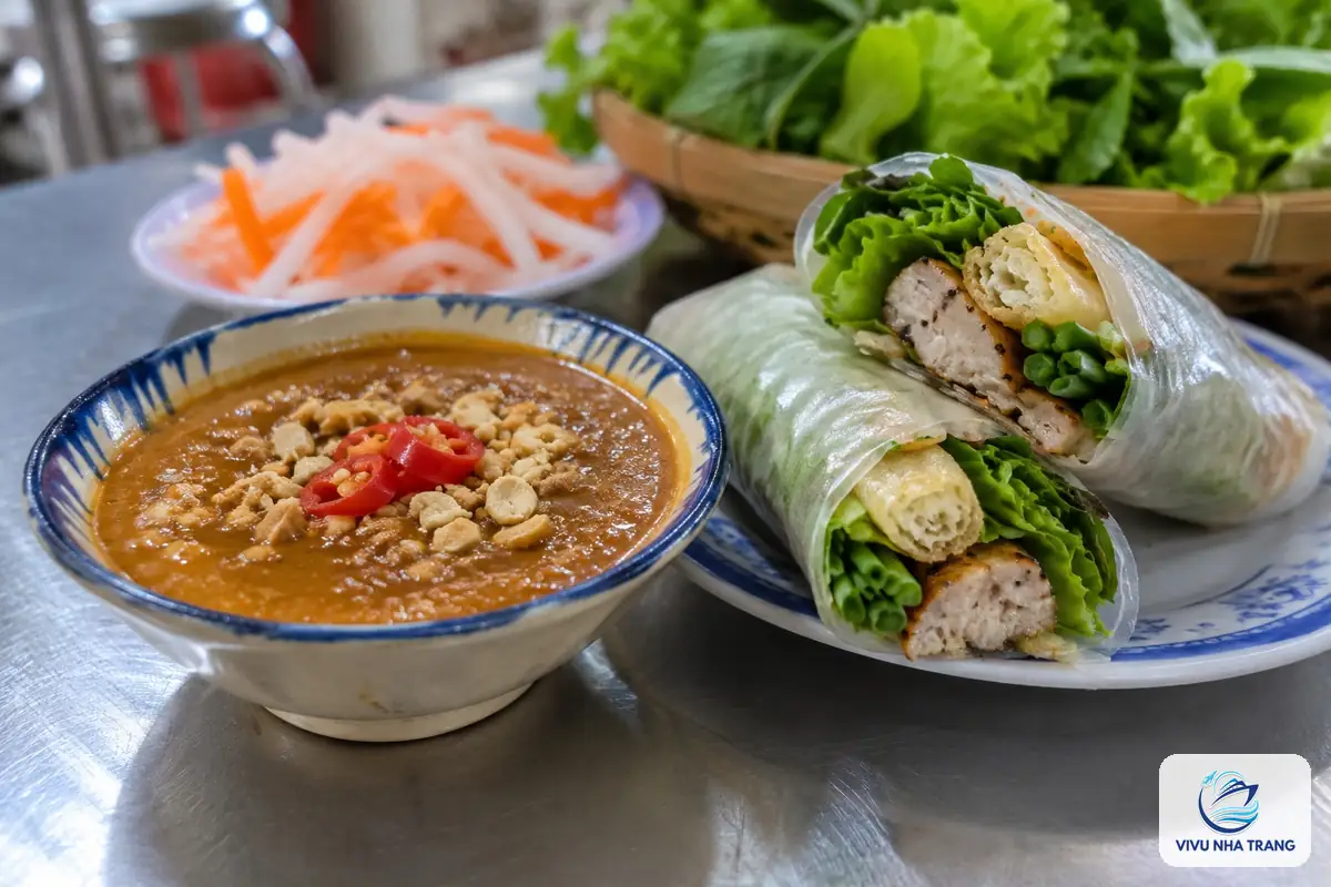 Chén nước chấm nem nướng Nha Trang màu cam nâu rắc đậu phộng, bên cạnh hai cuốn nem cắt đôi và rổ rau sống, ảnh minh hoạ